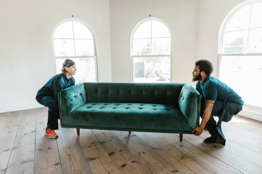 Two movers squatting to lift a dark-green velvet tufted sofa in a bright empty room with three arched windows and wide-plank wood floor