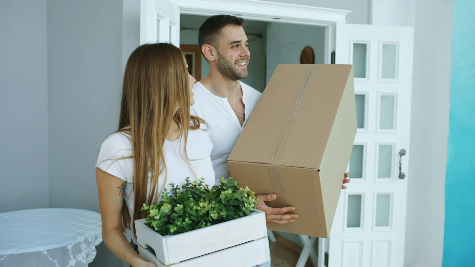 Smiling young couple in white t-shirts entering a new home: man holds a large cardboard box, woman holds a white crate of green plants