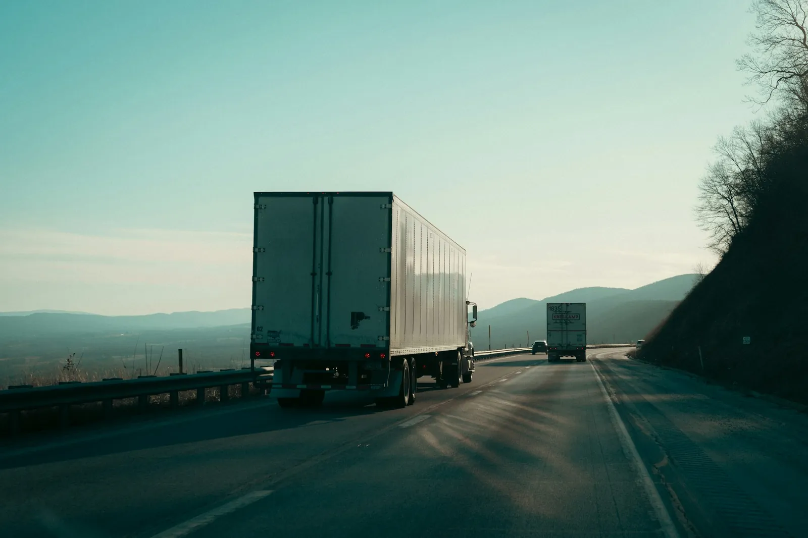Rear view of a plain white semi-trailer and a second truck heading down an interstate through hills, low sun, teal-toned sky