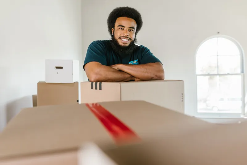 Friendly portrait of a smiling Black mover with afro and full beard leaning on stacked boxes, arms crossed, in a bright white room with arch