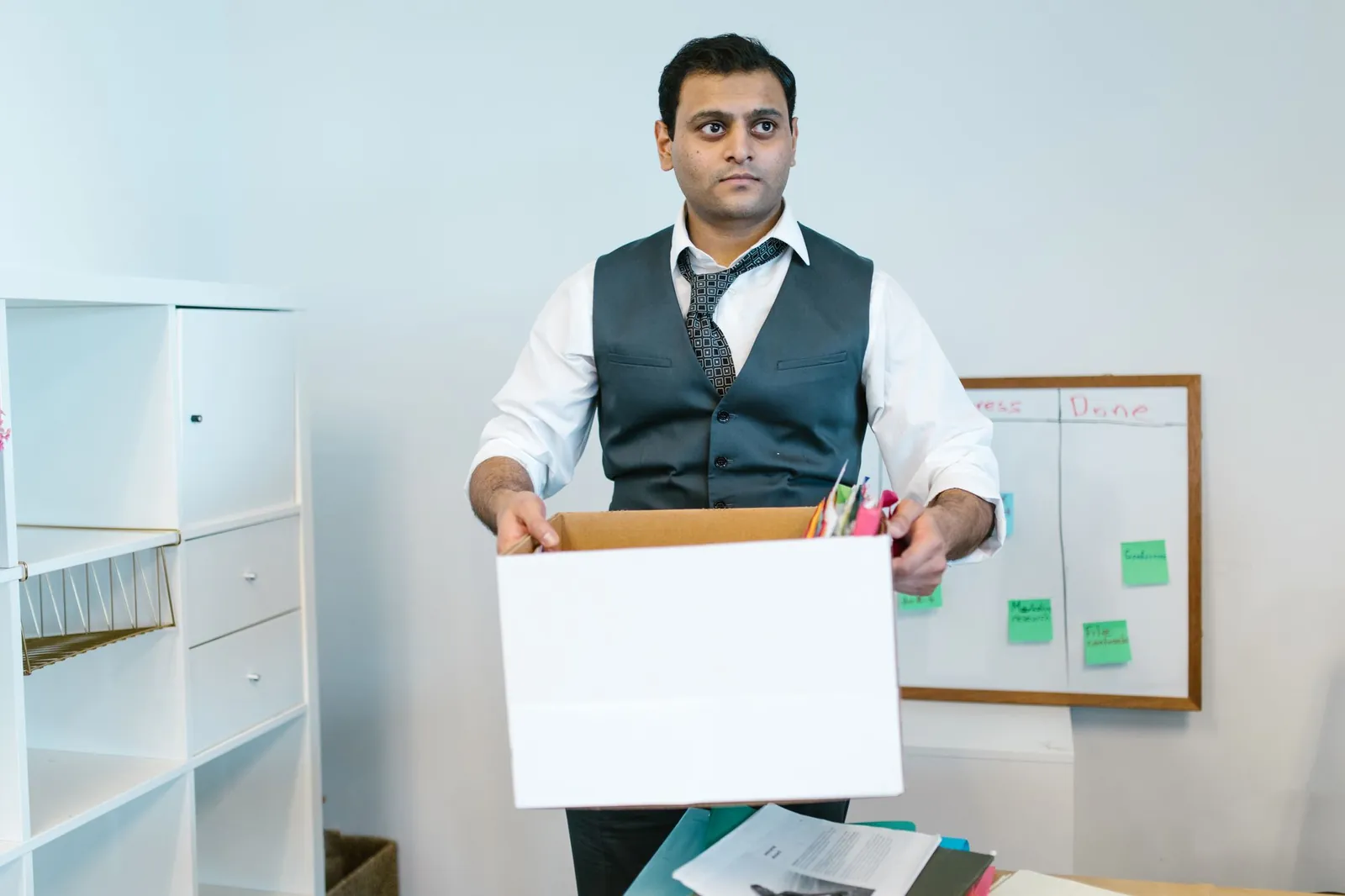South-Asian man in white shirt, loosened patterned tie and charcoal vest holding a white box of office belongings in a bright office with wh