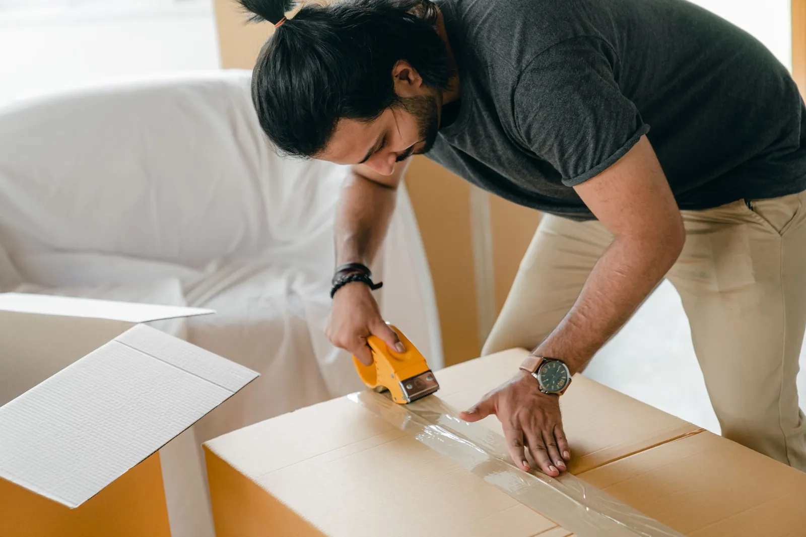 Man with long dark hair in a top-knot, dark charcoal t-shirt and khaki pants, sealing a cardboard box with an orange tape gun